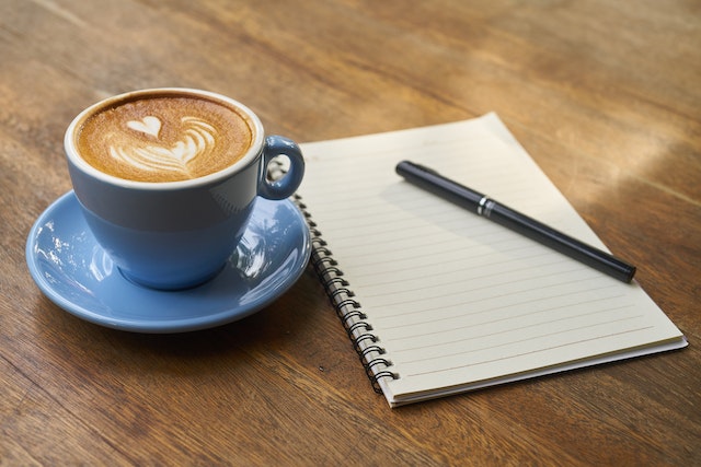 Latte in blue cup with matching saucer next to blank notepad and black pen